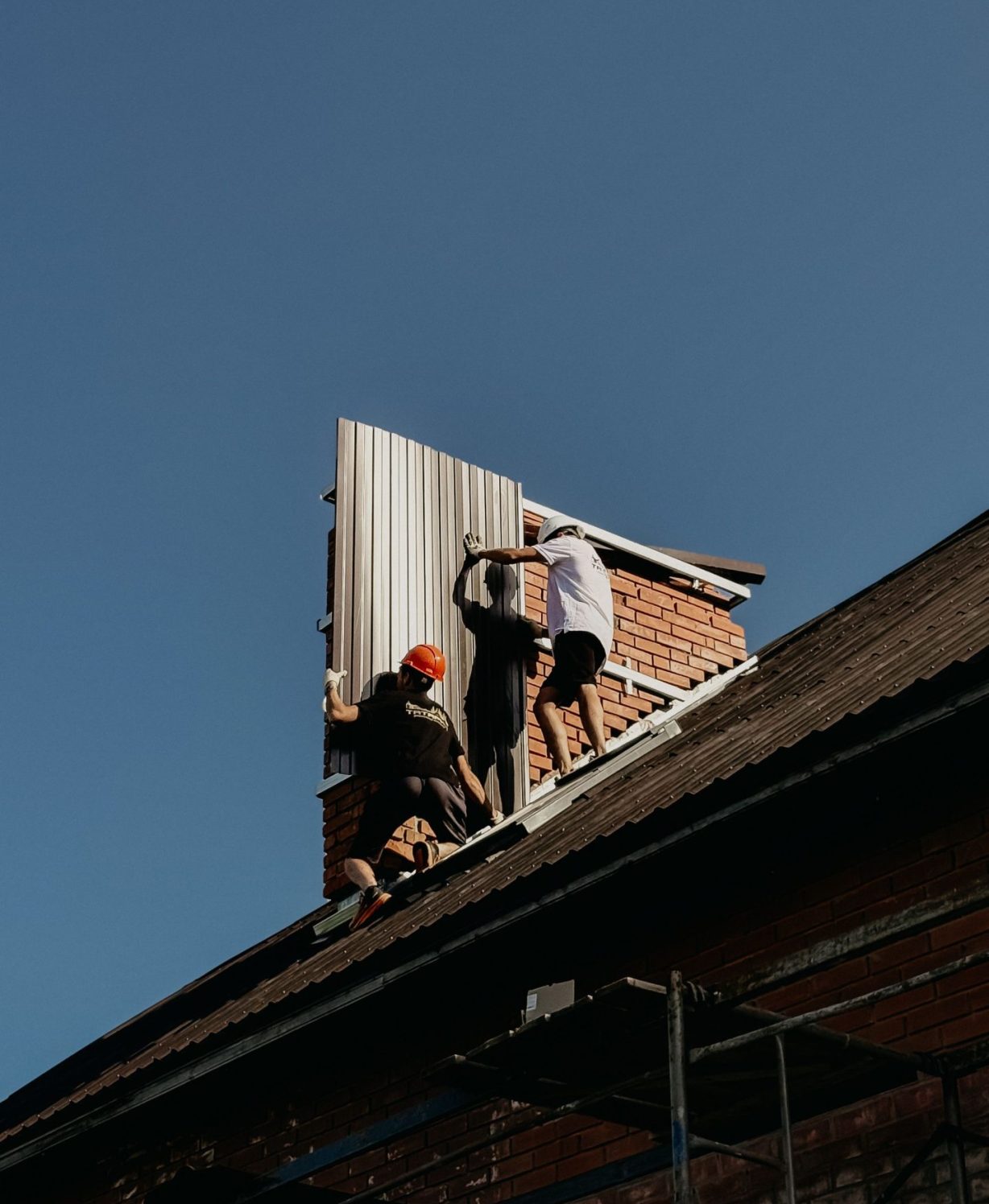 Three workers installing metal roof panels on a brick house under clear skies.
